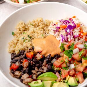 Angled view of a burrito bowl filled with quinoa, black beans, slaw, avocado, and pico de gallo, with a bowl of salsa in the background.
