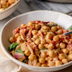 Creamy &ldquo;marry me&rdquo; chickpeas with sun-dried tomatoes and spinach served in a white bowl with a spoon.