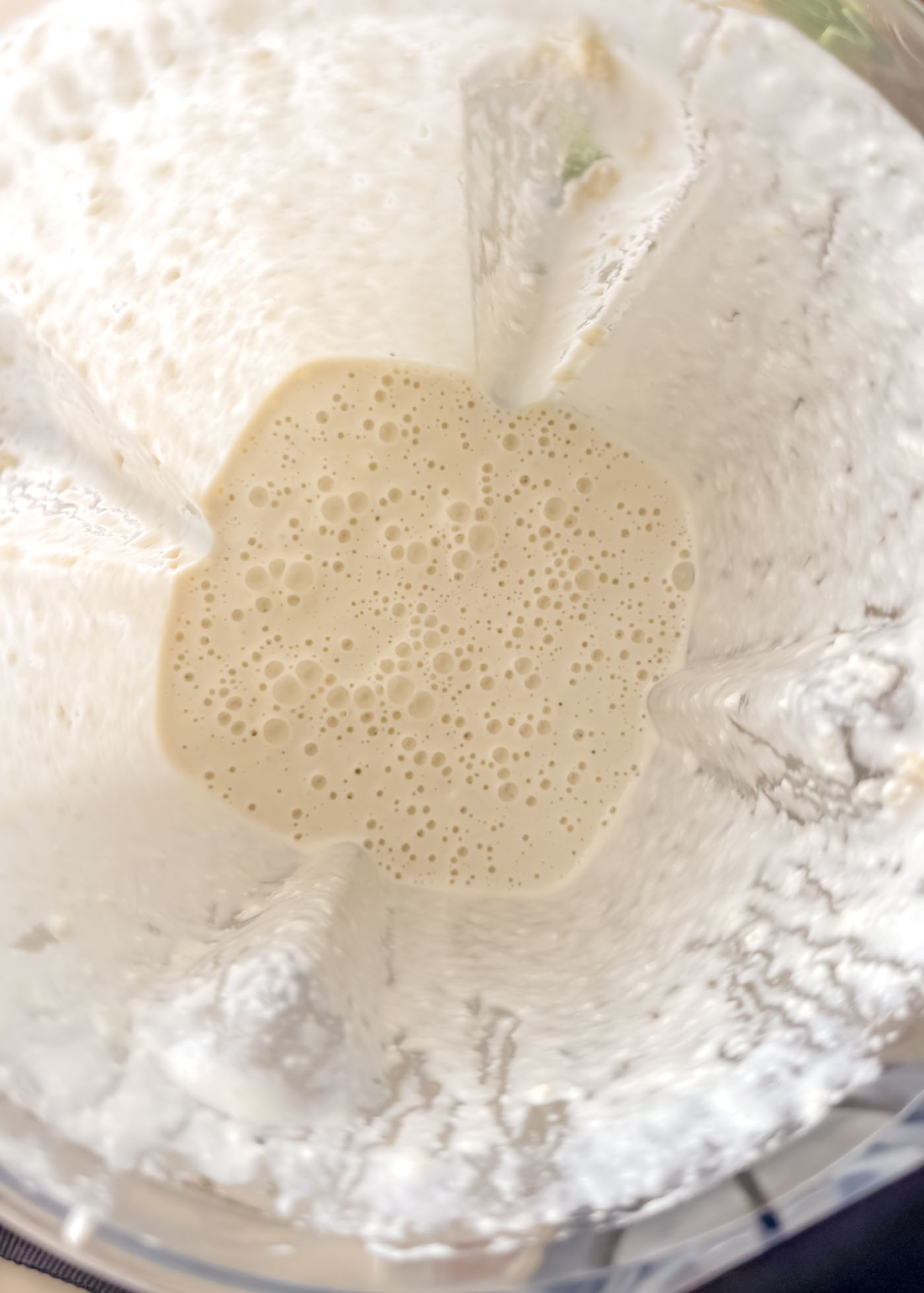 Top-down view inside a blender containing a smooth, pale cashew cream with small air bubbles after blending soaked cashews and broth.