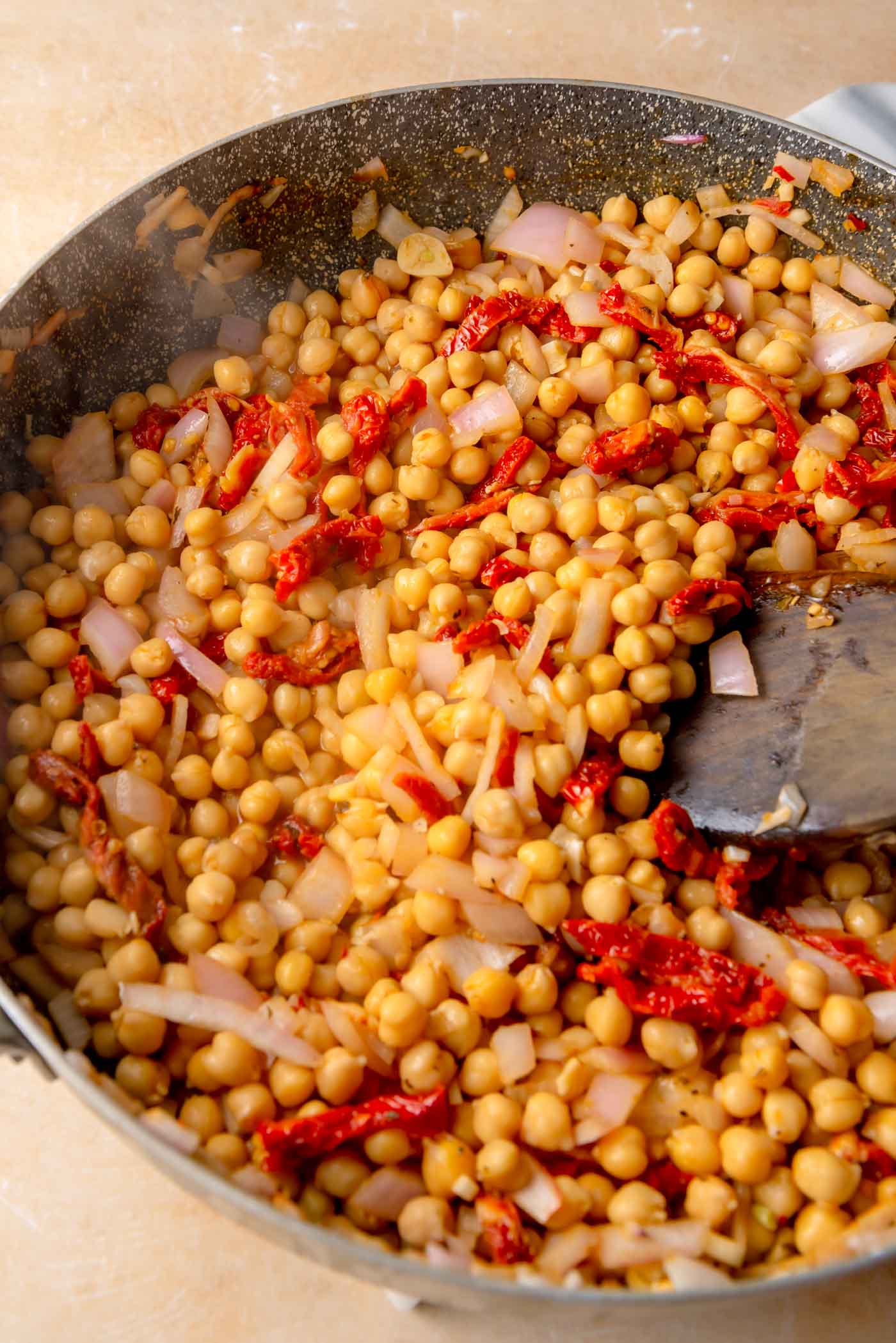 Cooked chickpeas and sliced sun-dried tomatoes in a skillet with an onion mixture, being stirred together as the flavors combine.