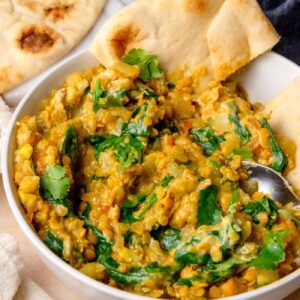 Bowl of spinach red lentil curry served with piece of naan and fresh cilantro on top.