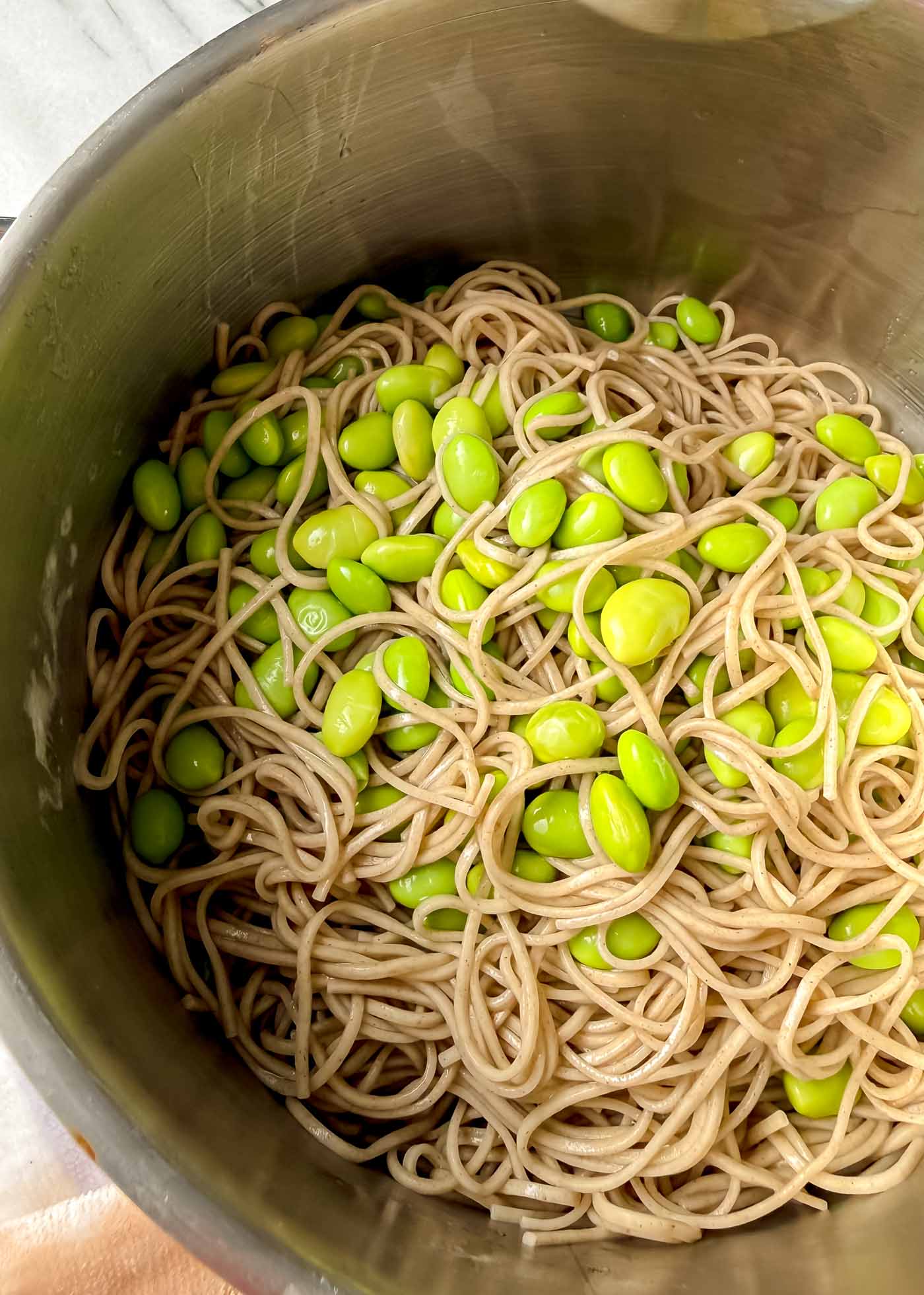 A stainless steel pot filled with cooked soba noodles tangled together with bright green edamame beans, viewed from above so the glossy noodles and beans fill the frame.