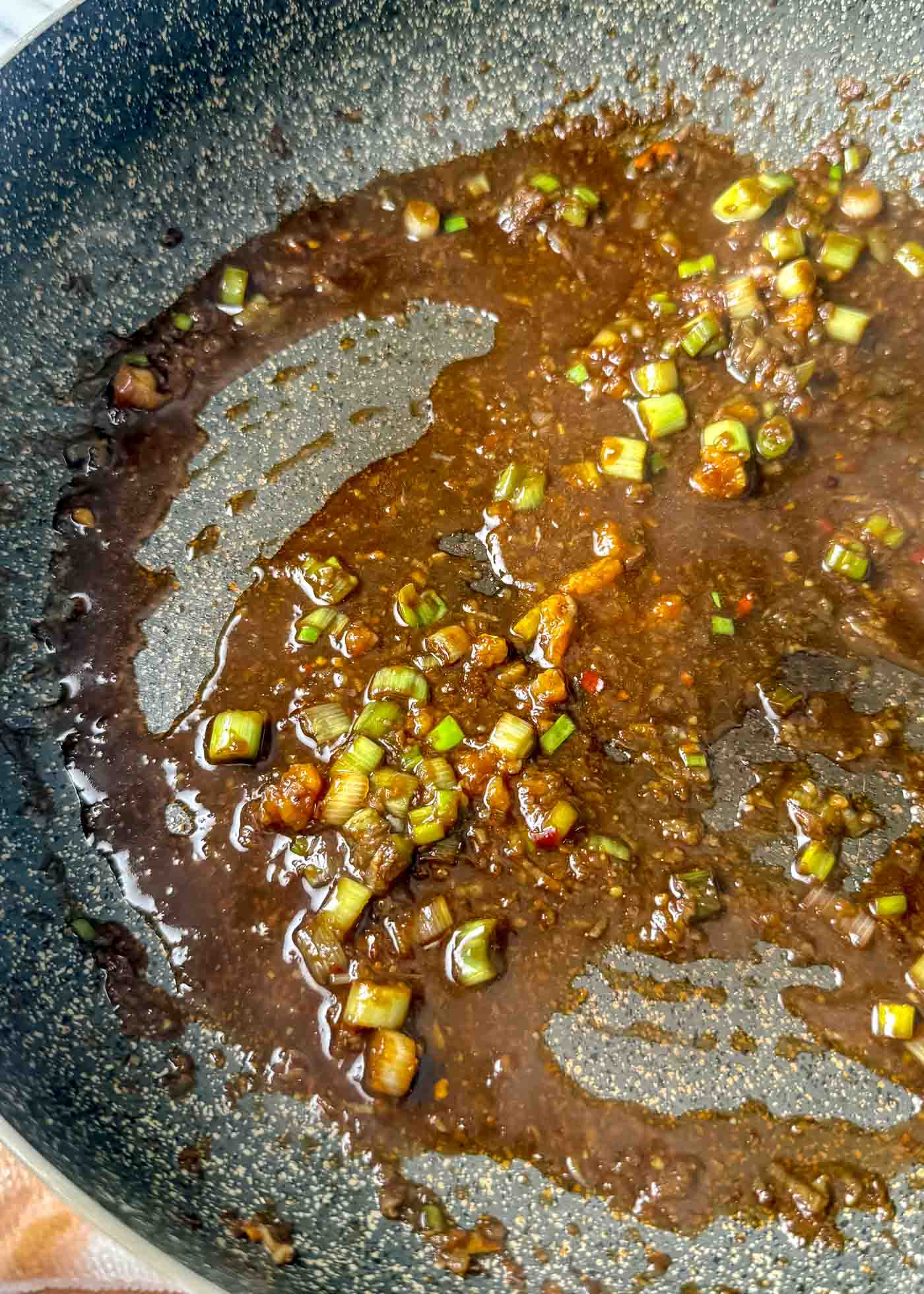 Dark glossy sesame-soy sauce simmering in a pan with chopped green onions, garlic, and small chili flakes.