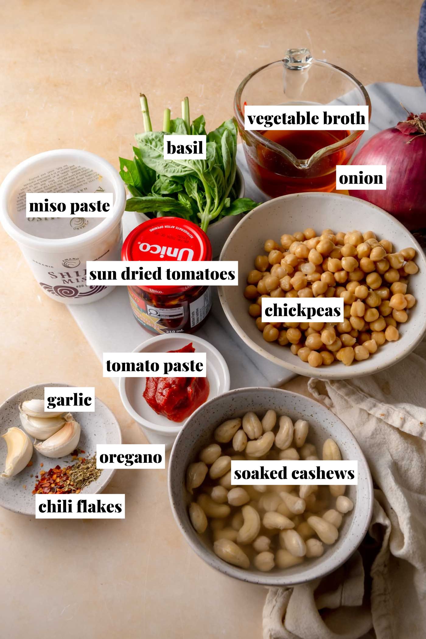 Overhead view of ingredients for creamy “marry me” chickpeas, including chickpeas, soaked cashews, sun-dried tomatoes, tomato paste, miso paste, vegetable broth, red onion, garlic, fresh basil, dried oregano, and chili flakes arranged on a countertop.