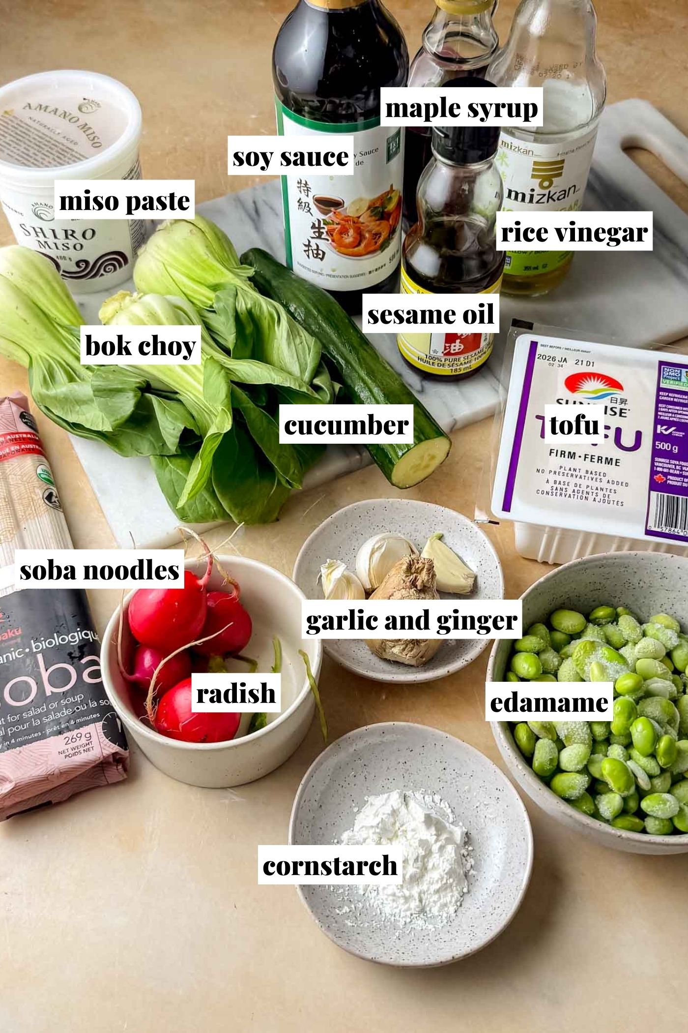 A countertop display of ingredients for tofu soba noodles, including bottles of soy sauce, maple syrup, rice vinegar, and sesame oil, a tub of white miso paste, fresh bok choy, a whole cucumber, a package of firm tofu, frozen edamame, dry soba noodles, red radishes, fresh garlic and ginger, and a small bowl of cornstarch arranged on a cutting board and in bowls.