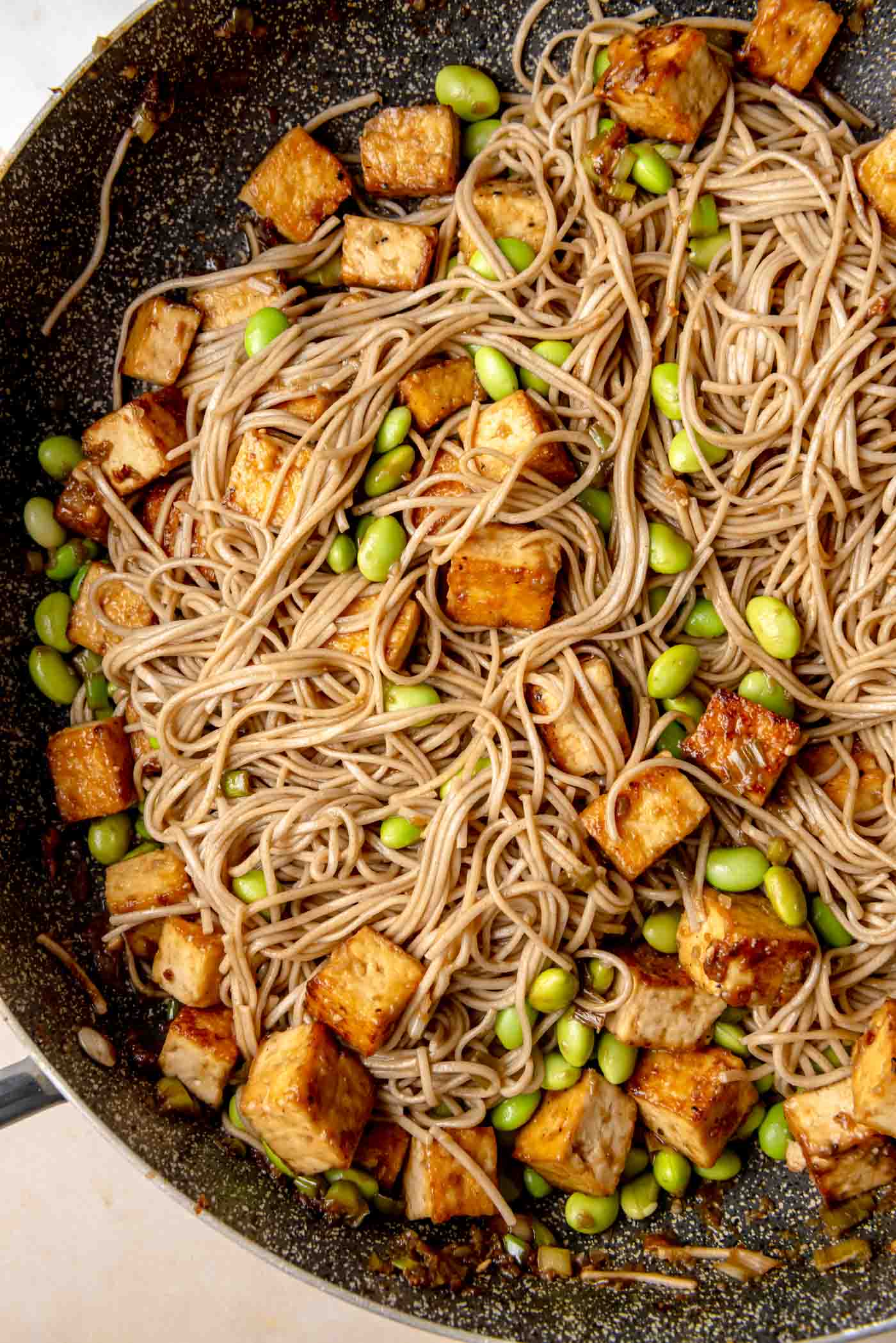 Soba noodles and crispy tofu cubes cooking together in a skillet with bright green edamame beans mixed throughout.