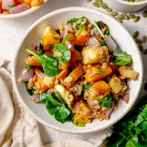 Overhead view of warm farro salad with roasted squash, apples, spinach, red onion and herbs, with pumpkin seeds and parsley nearby.