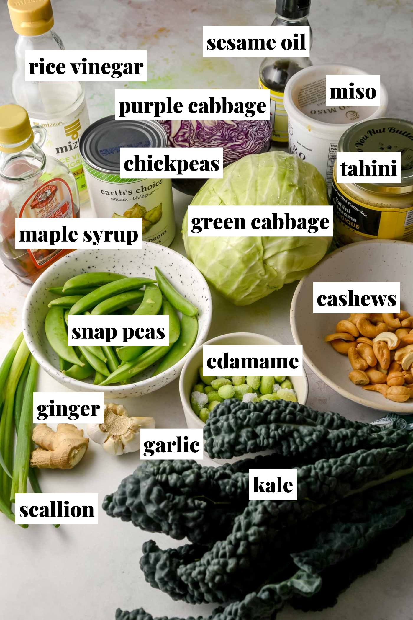 Miso crunch salad ingredients arranged on a countertop including kale, green cabbage, purple cabbage, chickpeas, edamame, snap peas, cashews, tahini, miso, sesame oil, rice vinegar, maple syrup, garlic, ginger and scallions. Each labelled with text overlay.