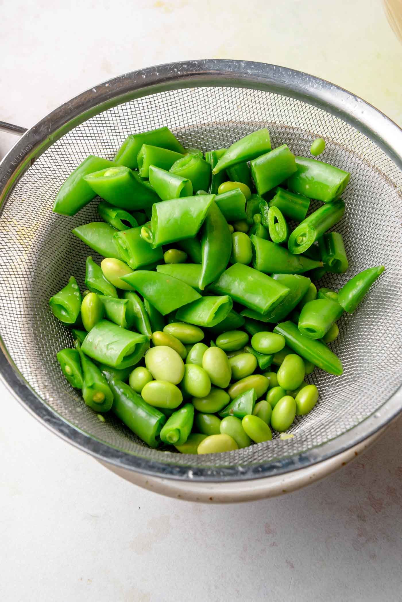 Fresh snap peas and shelled edamame draining in a mesh strainer, prepared for a miso crunch salad.