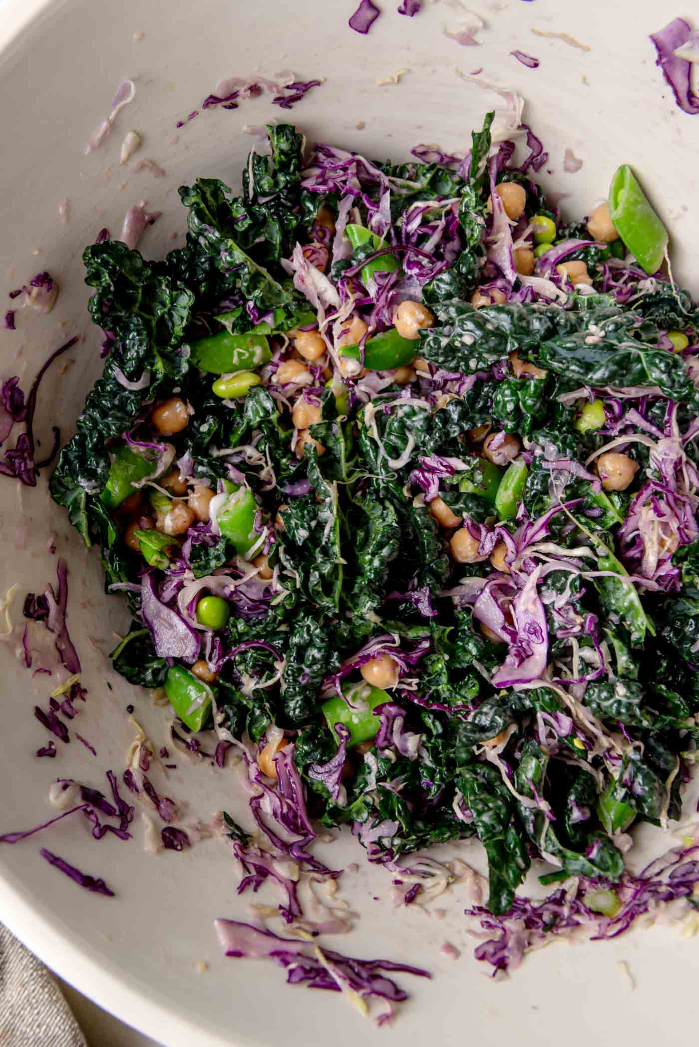Kale cabbage salad being tossed in a large bowl with chickpeas, edamame and snap peas coated in a creamy miso sesame dressing.