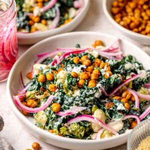 Roasted vegetable Caesar salad with crispy chickpeas, kale, broccoli, cauliflower, and pickled red onions in a creamy dressing, served in a shallow bowl.