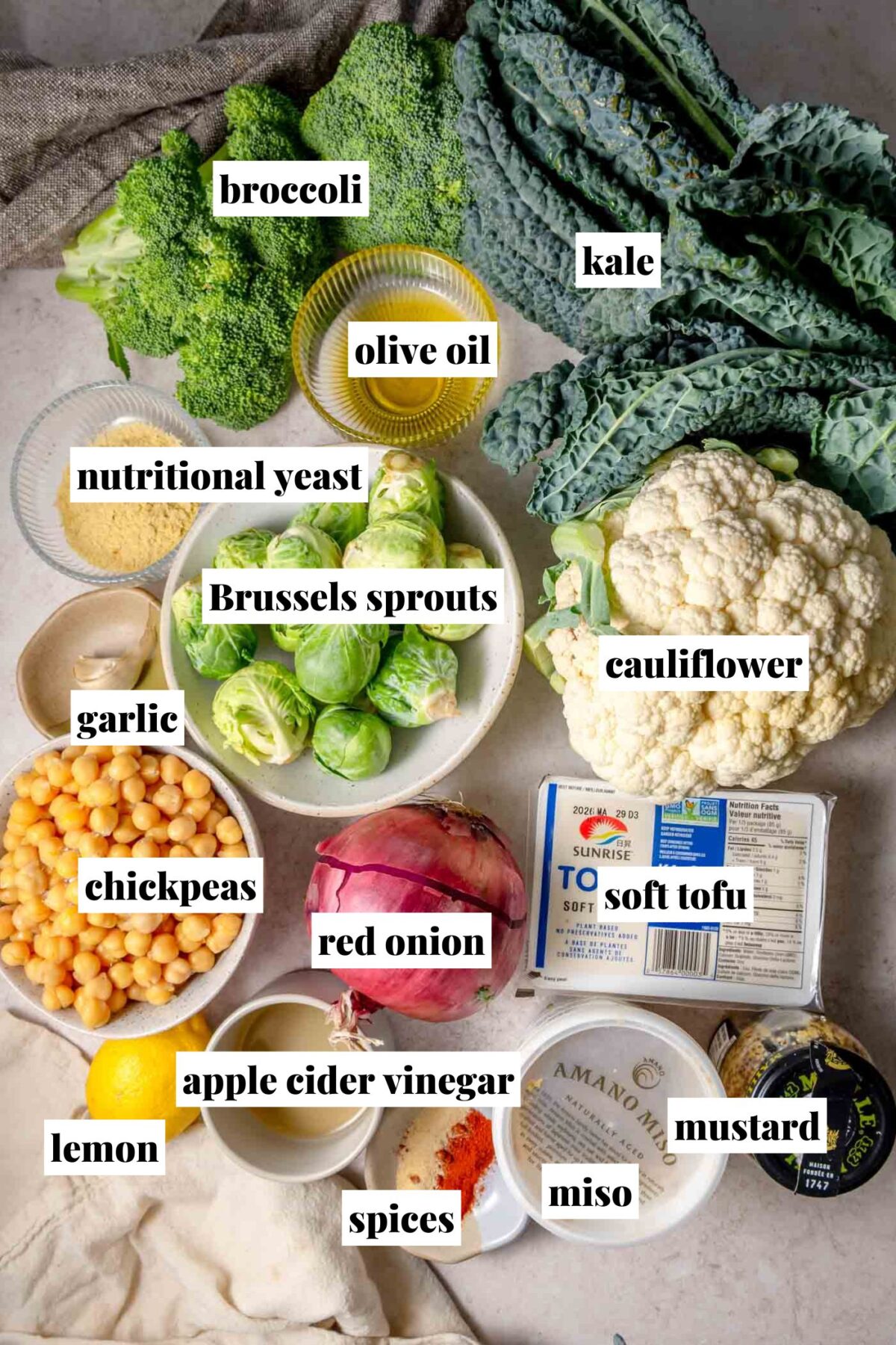 Overhead view of ingredients for roasted vegetable Caesar salad including broccoli, kale, cauliflower, Brussels sprouts, chickpeas, red onion, garlic, lemon, soft tofu, miso, mustard, apple cider vinegar, olive oil, nutritional yeast, and spices.