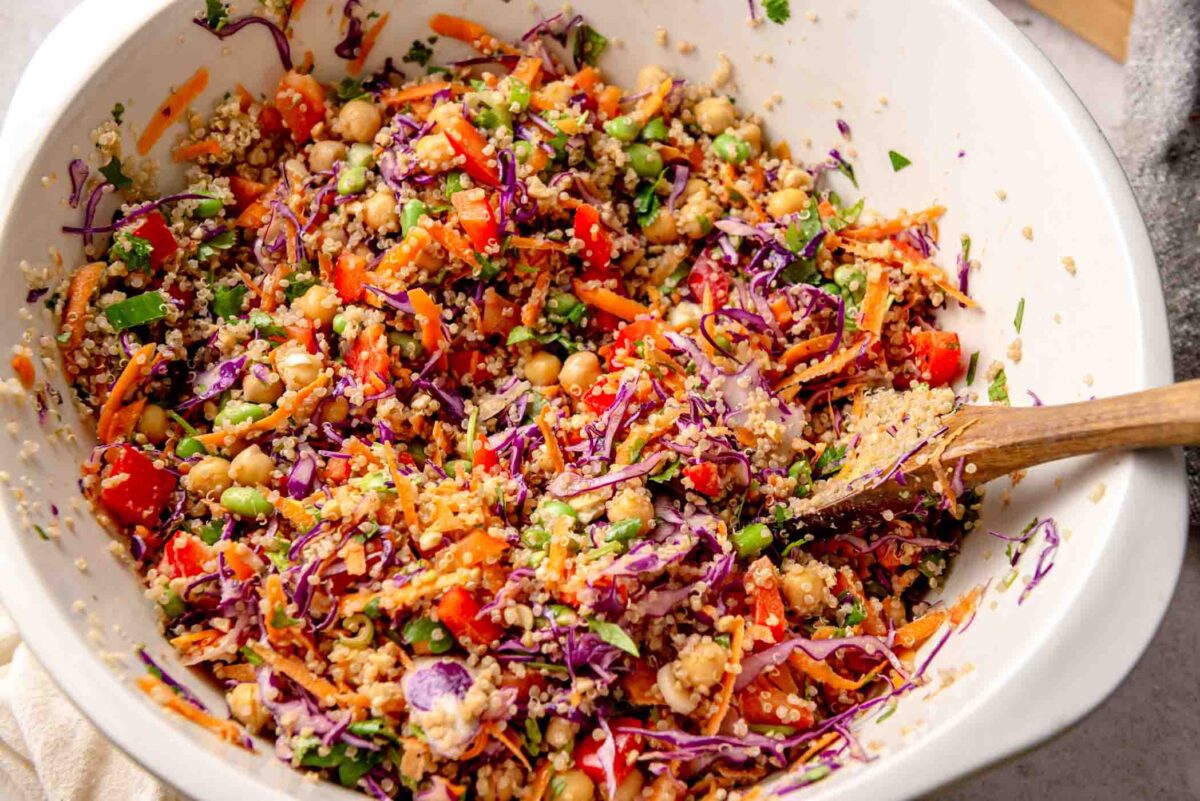 Thai peanut quinoa salad being tossed in a large bowl with shredded red cabbage, carrots, bell pepper, chickpeas, and herbs.