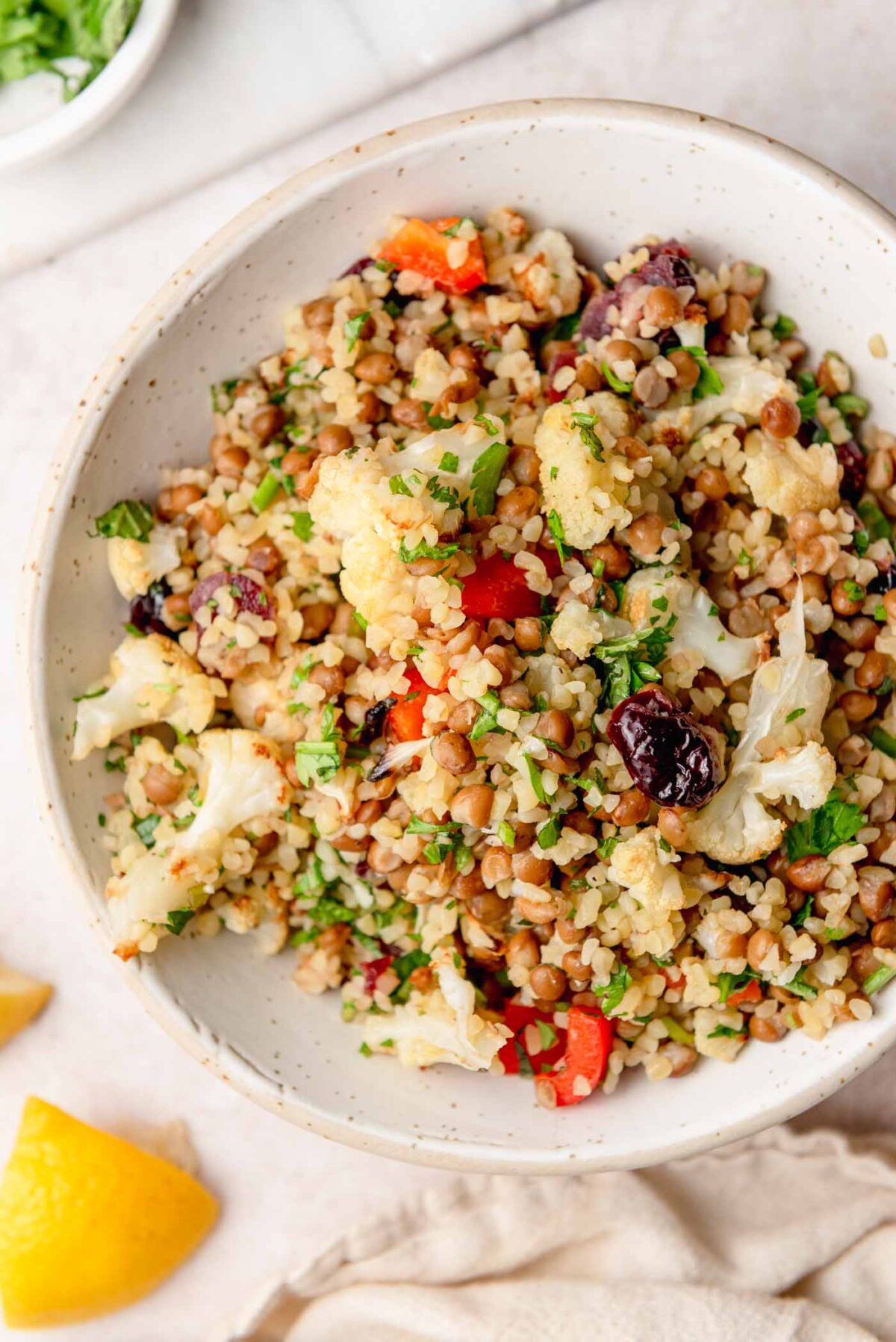 Bulgur salad with lentils, roasted cauliflower, red bell pepper, dried cranberries, parsley and mint, served in a white bowl.