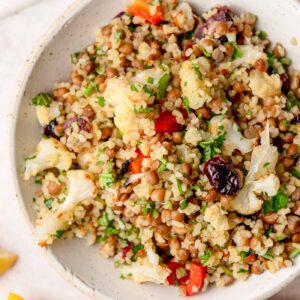 Bulgur salad with lentils, roasted cauliflower, red bell pepper, dried cranberries, parsley and mint, served in a white bowl.