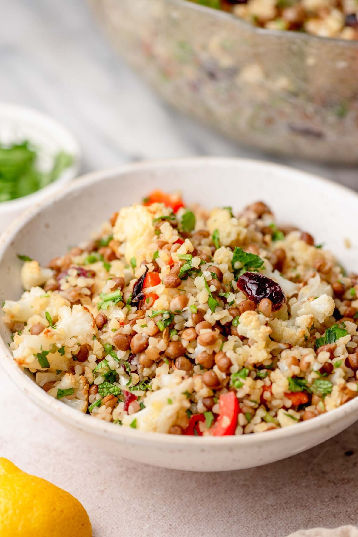 Bulgur salad with lentils, roasted cauliflower, red bell pepper, dried cranberries and fresh herbs served in a white bowl with a lemon wedge.