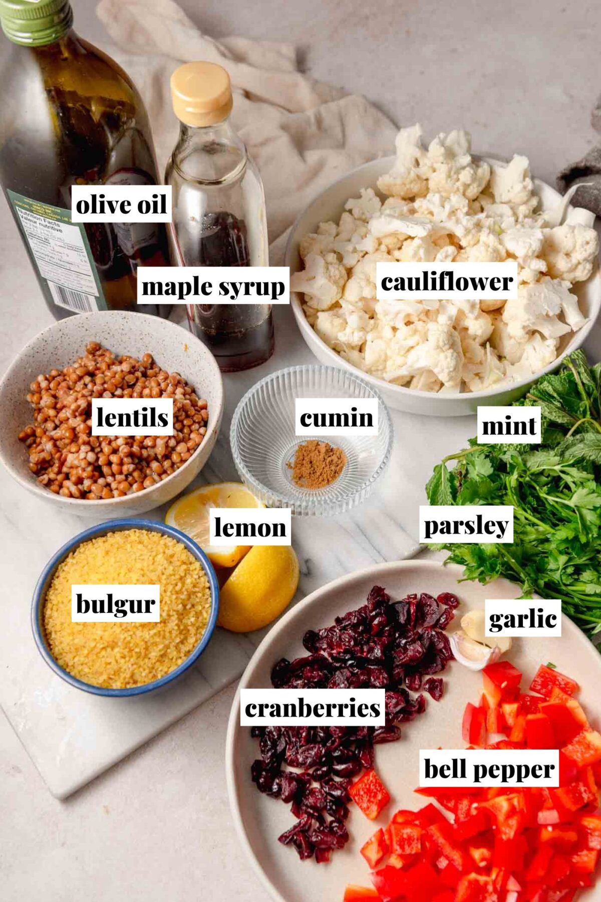 Ingredients for bulgur salad including bulgur, lentils, cauliflower, red bell pepper, dried cranberries, parsley, mint, lemon, garlic, cumin, maple syrup and olive oil, labeled in the image.